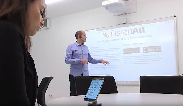 En un aula un profesor que es Jose Maria Fernandez Gil explica sobre una proyección y lleva un micrófono de solapa mientras una estudiante está sentado y está viendo en su móvil el texto que se va reconociendo en ListenAll